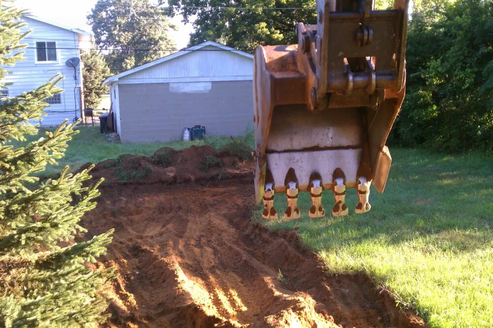 Excavator bucket cutting a driveway beside a home